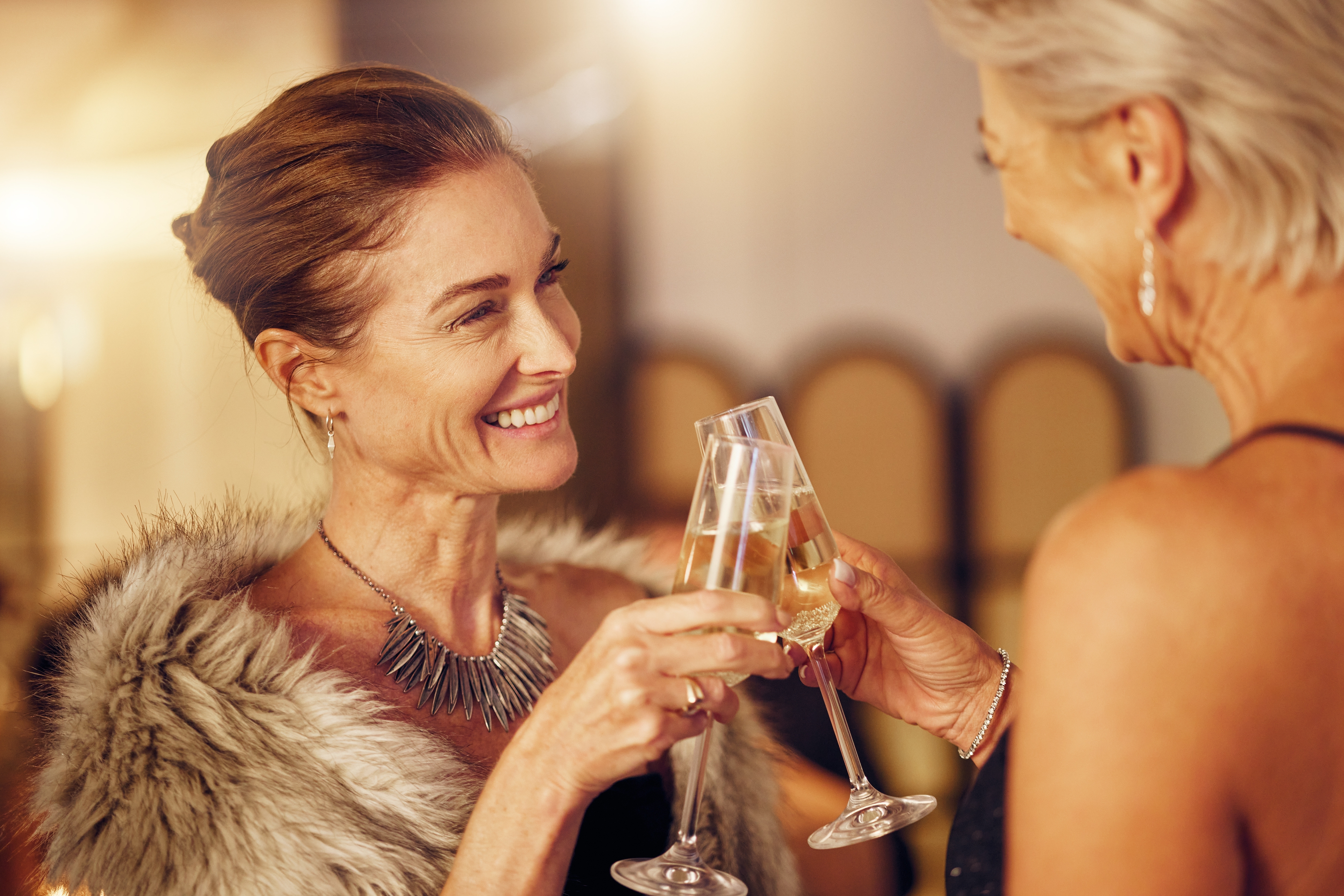 A pair of middle aged woman clink champagne glasses together.