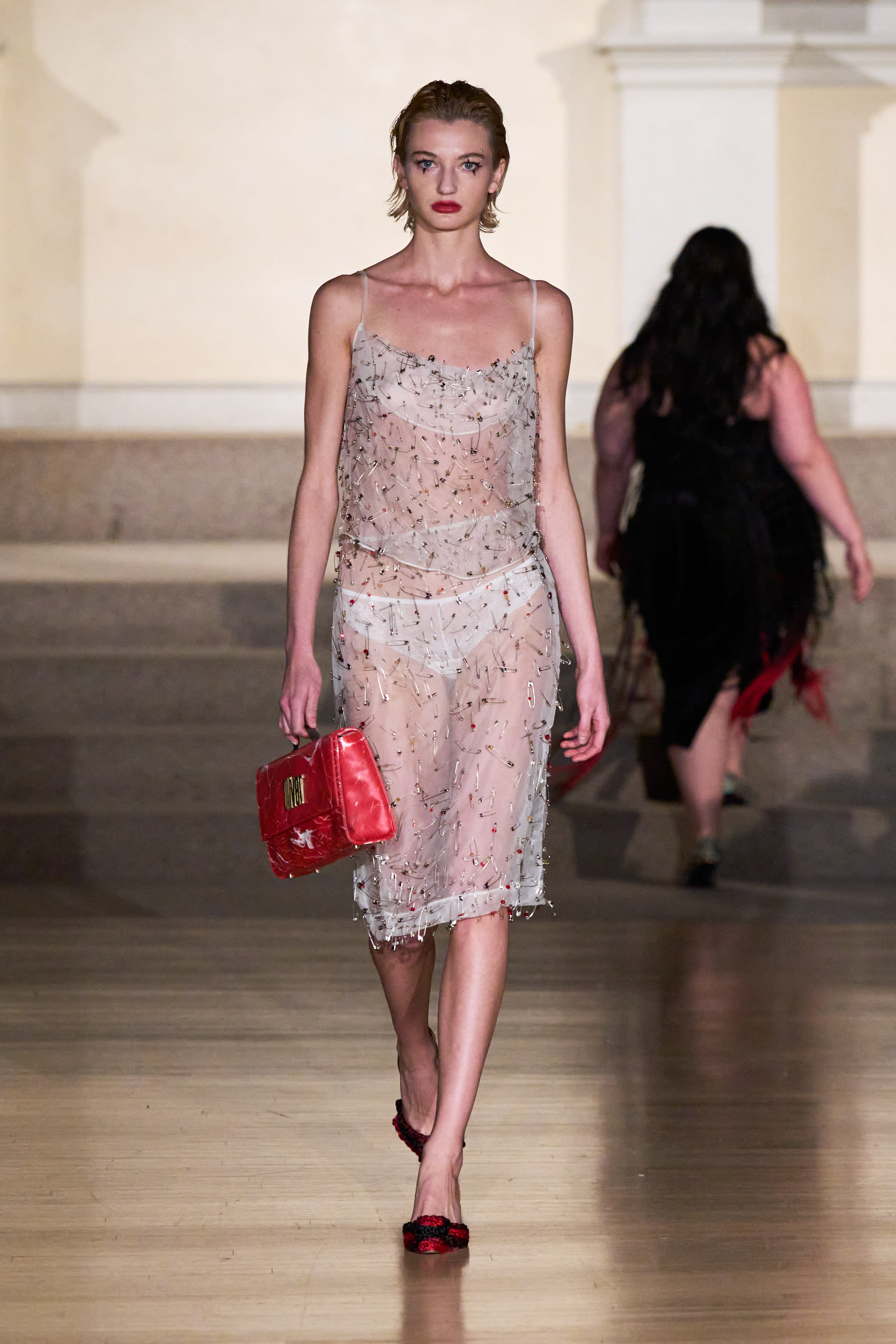 a woman walking down a runway wearing a sheer dress with commonly found objects like safety pins and forks on it.