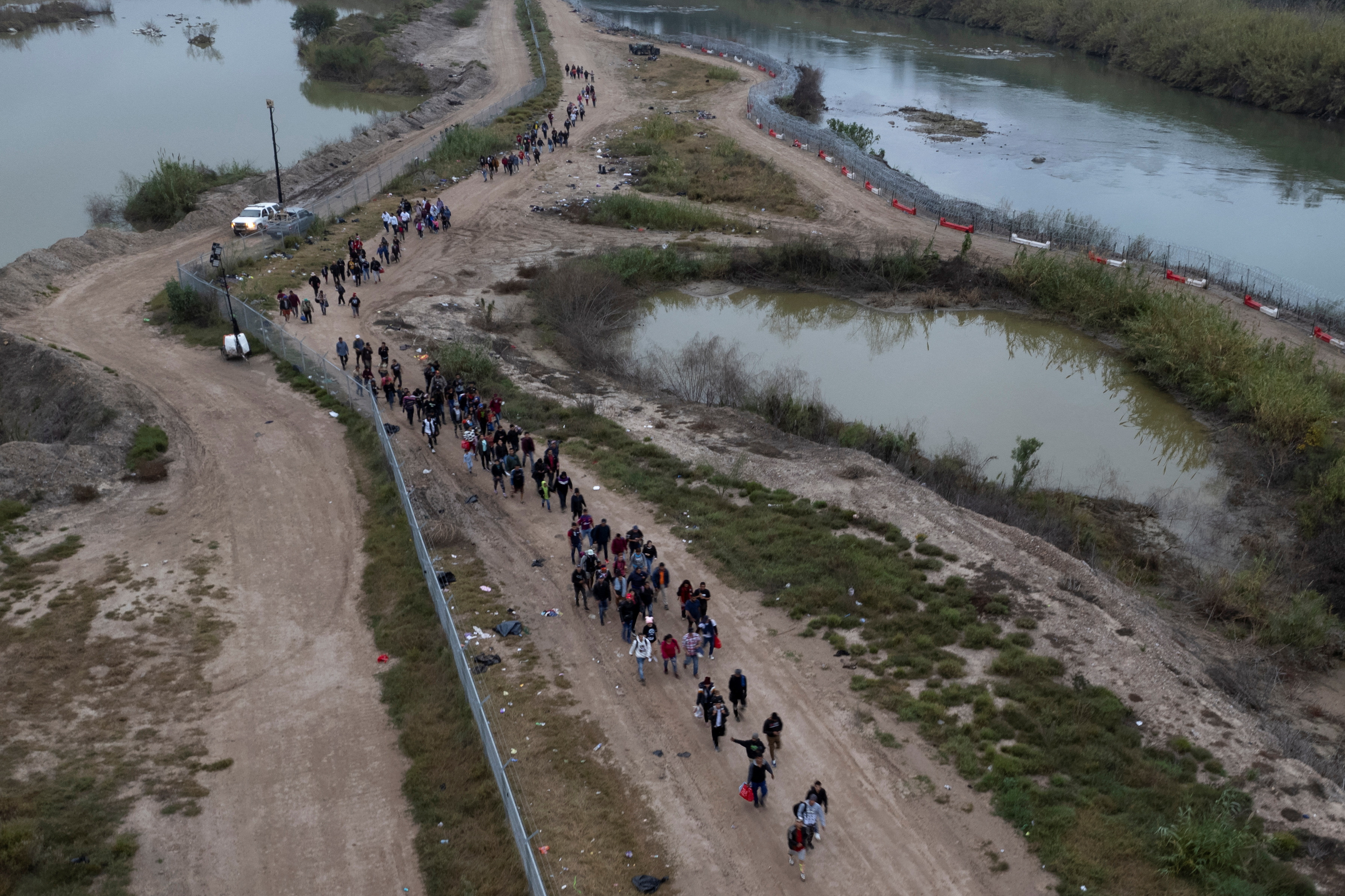 Migrants wade across Rio Grande River, walk on dirt road towards a processing center, Eagle Pass, Texas.