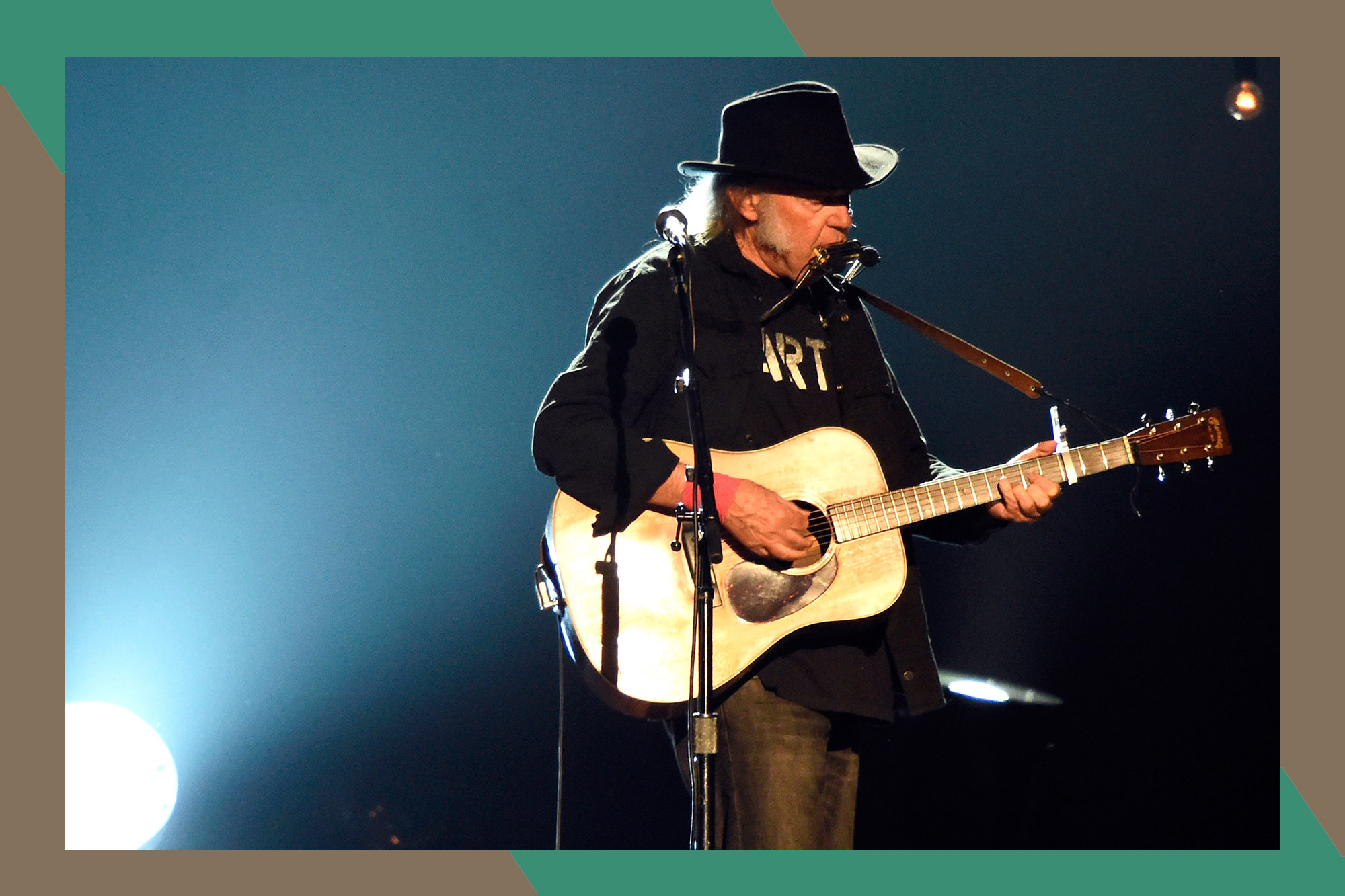 Neil Young plays guitar and harmonica while wearing a cowboy hat.